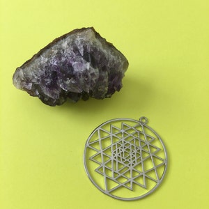 May include: A purple amethyst crystal cluster and a silver metal pendant with a geometric design. The pendant features a circular shape with a complex pattern of triangles and lines.