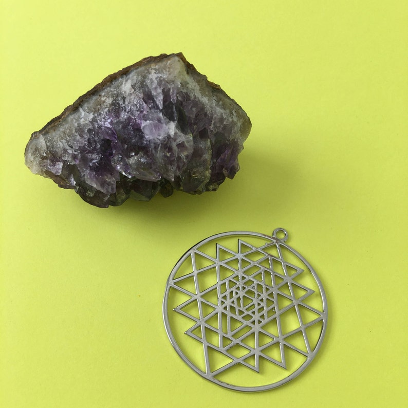 May include: A purple amethyst crystal cluster and a silver metal pendant with a geometric design. The pendant features a circular shape with a complex pattern of triangles and lines.