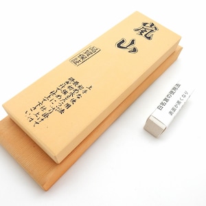 Puede incluir: Una piedra de afilar de color marrón claro con texto japonés en la parte superior y lateral. La piedra está colocada sobre una base de madera. Una pequeña caja blanca con texto japonés se encuentra en la esquina inferior derecha de la imagen.