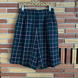 May include: High-waisted shorts featuring a dark green and navy blue plaid pattern with thin white grid lines. The shorts include a button closure, a waistband with belt loops, and subtle front pleats, hanging from a gold-toned metal hanger.