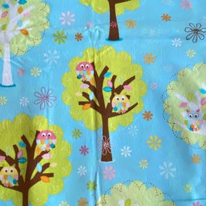 May include: A blue fabric with a repeating pattern of green trees with brown trunks and colorful owls. The owls are perched on the branches of the trees. There are also small, colorful flowers scattered throughout the fabric.