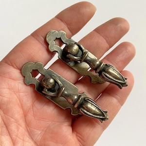May include: Two antique-style metal drawer pulls with a tarnished silver finish. Each pull features a rectangular backplate with scalloped edges and a hinged, teardrop-shaped handle that hangs downward from a central rounded mounting point.