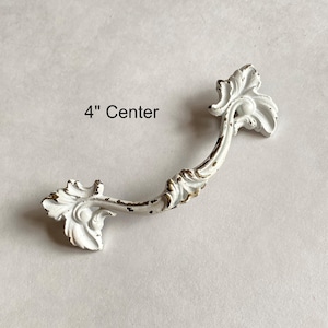 May include: White and gold distressed cabinet pull with ornate floral detailing. The pull measures 4 inches (10.16 cm) center to center, featuring a curved handle and decorative ends. The text "4" Center" is visible.