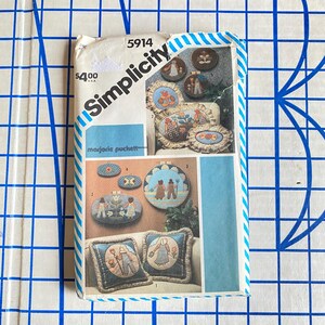 May include: A vintage Simplicity pattern envelope featuring embroidered pillow and wall hanging designs. The envelope displays various finished projects with detailed illustrations, including floral and figure motifs. The price is $4.00.
