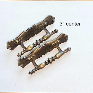 May include: Two antique brass drawer pulls with a 7.6 cm centre measurement. The pulls have a decorative design with a handle and ornate detailing. The hardware is a warm, aged brass colour.