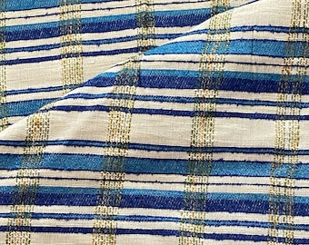 May include: Close-up of a woven fabric with a plaid pattern. The fabric features alternating stripes of light beige, blue, and dark blue. The texture appears slightly rough, with a mix of colors and a subtle sheen. The fabric is folded over itself.