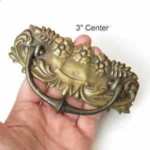 May include: Antique brass drawer pull with a floral design and a 3" centre to centre measurement. The pull has a curved handle and ornate details.
