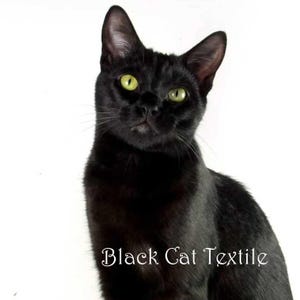 以下が含まれることがあります： カメラに向かってまっすぐ見つめる緑色の目をした黒い猫。猫は白い背景に座っています。猫の下に「Black Cat Textile」という文字が書かれています。