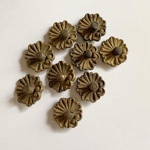 May include: A collection of nine circular brass buttons, each featuring a raised floral petal design radiating from a central spiral knob. The metal has a muted, aged finish with detailed grooves and scalloped edges on every individual piece.