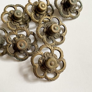May include: A collection of antique-style brass drawer knobs featuring a circular, open-work floral design with a central raised button. Each knob has a weathered, tarnished finish with visible patina, showcasing intricate petal-like metal filigree.