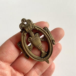 May include: An oval-shaped brass drawer pull featuring a central urn motif suspended within a decorative frame. The metal hardware has a weathered, aged finish with intricate detailing along the outer border and a small mounting hole at the top.