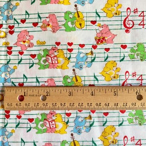 May include: White fabric with a repeating pattern of Care Bears, musical notes, and red hearts. The Care Bears are in various colors and poses. A wooden ruler with inch measurements is at the bottom.