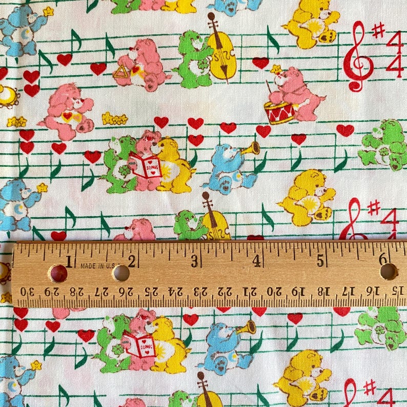 May include: White fabric with a repeating pattern of Care Bears, musical notes, and red hearts. The Care Bears are in various colors and poses. A wooden ruler with inch measurements is at the bottom.