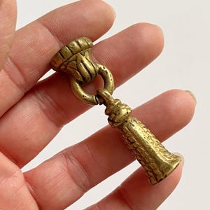 May include: A metallic gold-toned brass drawer pull featuring a decorative tassel design suspended from a circular ring. The hardware has a textured, aged finish with a flared top mounting piece and a detailed, cylindrical tassel drop component.