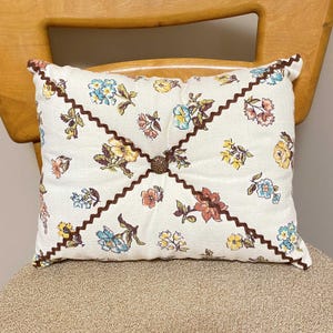 May include: A white pillow with a brown floral pattern and brown scalloped trim. The pillow has a button closure in the center.