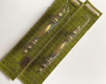 May include: Two antique brass finish cabinet pulls with a decorative, multi-tiered spindle design. Each metal handle is securely encased in clear plastic packaging attached to a green cardboard backing featuring the brand name Colonnade.