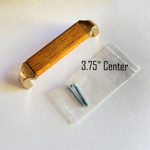 May include: A cabinet pull with a light brown wooden center and gold-colored metal ends. The text "3.75" Center" is visible. Includes a small bag with two screws.