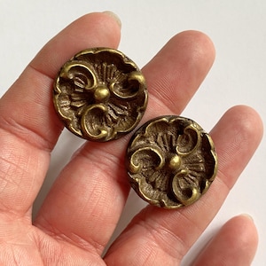 May include: Two circular brass-toned buttons featuring a raised, ornate floral-inspired relief pattern. Each button has a central circular point surrounded by radiating lines and curved, scroll-like details along the outer edge of the face.