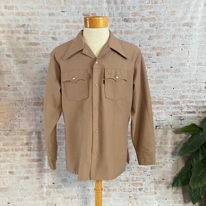 May include: A tan button-down shirt with long sleeves, two chest pockets, and a pointed collar. The shirt is displayed on a mannequin and has decorative stitching details. The shirt is a neutral color and has a classic design.
