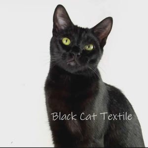 May include: A sleek black cat with bright green eyes is the focal point of this image. The cat is positioned against a plain white background. The text "Black Cat Textile" is written in a cursive font across the bottom of the image.