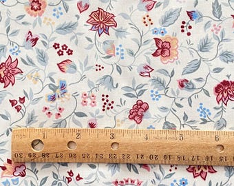 May include: A white fabric with a repeating floral pattern. The pattern features small pink, red, and blue flowers with green leaves. A wooden ruler is placed on the fabric for scale.