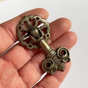 May include: A brass-toned metal furniture pull featuring a circular, openwork backplate with a scalloped edge and a hinged, drop-style handle. The handle has a decorative, symmetrical base with curved, scroll-like details and a central vertical column.