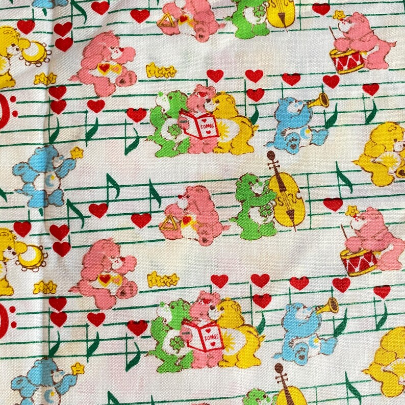 May include: White fabric with a repeating pattern of Care Bears, musical notes, and red hearts. The Care Bears are in various colors, including pink, yellow, blue, and green. Some bears are playing musical instruments.