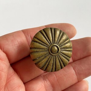 May include: A round, brass-toned metal cabinet knob featuring a sunburst pattern with raised ridges radiating from a central circular point. The surface has a weathered, matte finish with subtle variations in color across the textured metal.