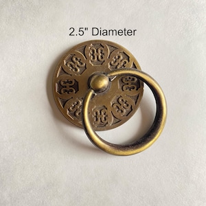 May include: Antique brass-toned cabinet pull with a circular design, featuring an ornate pattern and a ring handle. The diameter is 2.5 inches. This decorative hardware adds a vintage touch to furniture and cabinetry.