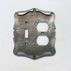 May include: A decorative, textured metal light switch plate with a single toggle switch and two electrical outlets. The plate has a unique, ornate border and a mottled, gray finish, adding a rustic touch to any room. The plate is designed to fit standard electrical boxes.
