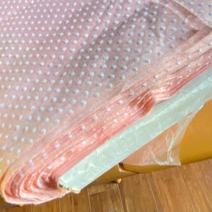 May include: A stack of folded pink fabric with a white polka dot pattern. The fabric is wrapped in clear plastic and rests on a wooden surface. The fabric has a textured appearance.