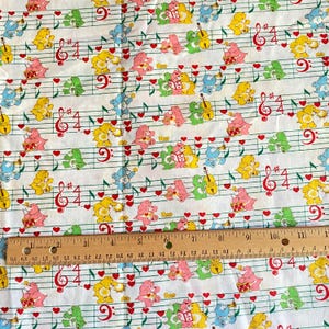 May include: White fabric with a repeating pattern of colorful cartoon bears, musical notes, and red hearts. The bears are in various poses, playing instruments or reading books. A ruler is placed across the bottom of the fabric.