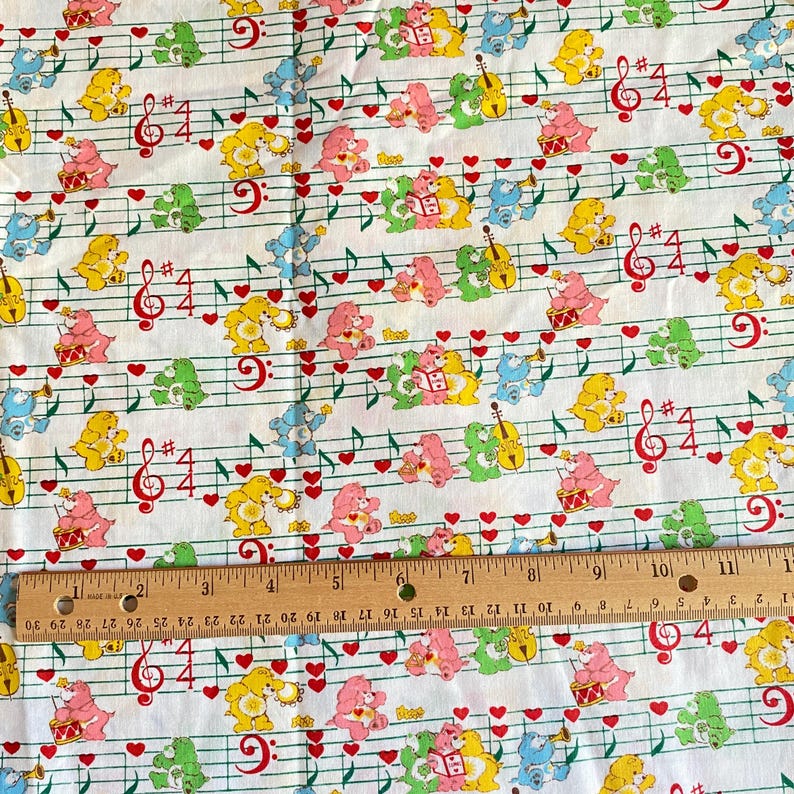 May include: White fabric with a repeating pattern of colorful cartoon bears, musical notes, and red hearts. The bears are in various poses, playing instruments or reading books. A ruler is placed across the bottom of the fabric.