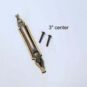 Peut inclure: Une poignée de meuble de couleur bronze avec un entraxe de 7,6 cm, accompagnée de deux vis. La poignée présente un design rectangulaire avec des détails ornés et un panneau central texturé, idéale pour la restauration de meubles.