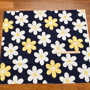 May include: Navy blue fabric featuring a repeating pattern of large white and yellow daisies. The daisies have white petals and yellow centres. A wooden ruler is placed on the fabric.
