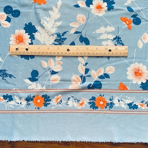 May include: Blue cotton fabric with a white and orange floral print. The fabric has a subtle texture and a border with a repeating pattern of orange flowers and leaves.