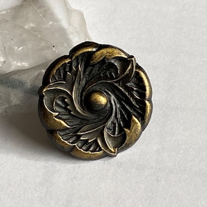 May include: A decorative, antique-style cabinet knob. The knob is round with a detailed floral design in shades of bronze and gold. The center features a small, raised sphere. The knob is likely made of metal.