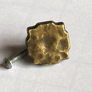 May include: A close-up of a brass-colored cabinet knob with a textured, irregular surface. The knob is square with rounded edges and is attached to a silver screw. The hardware is likely for a drawer or cabinet.