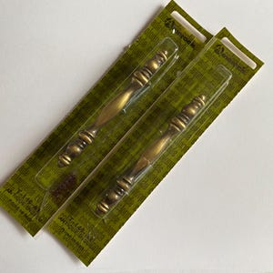 May include: Two antique brass-finished metal cabinet pulls with a decorative, multi-segmented spindle design. Each handle is individually packaged in a clear plastic blister pack attached to a green cardboard backing card labeled Colonnade.