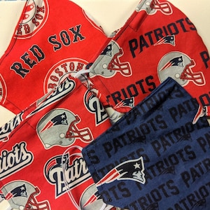 May include: Three fabric face masks with different sports team logos. The first mask is red with white text that says "Red Sox" and a white logo of a baseball cap. The second mask is red with white text that says "Patriots" and a white logo of a football helmet. The third mask is blue with white text that says "Patriots" and a white logo of a football helmet.