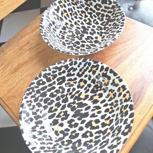 May include: Two ceramic bowls with a leopard print design. The bowls have a white background with black and tan spots. One bowl is round and the other is a shallow bowl. The bowls are on a wooden surface.