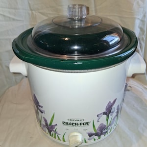 May include: A white Rival Crock-Pot stoneware slow cooker with a dark green rim and a clear glass lid. The exterior features a purple iris floral pattern, white side handles, and a front control knob with Off, Low, and High settings.
