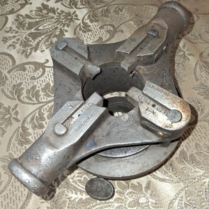May include: A vintage, industrial-style tool with a circular base and three adjustable arms. The metal tool has a worn, aged appearance with visible text, including "H.B. JARDINE & CO" and "HESHELEFRONT". The tool is likely used for gripping or holding objects.