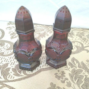 May include: A pair of red glass salt and pepper shakers with a decorative design. The shakers have a rounded body and a pointed top.