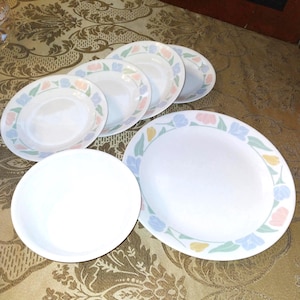 Peut inclure: Ensemble de six assiettes en céramique blanche avec un motif floral dans des tons de rose, bleu et vert. Les assiettes sont disposées en cercle sur une nappe à motifs dorés.