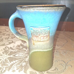 May include: A handmade ceramic mug with a unique shape and a blue and green glaze. The mug features a handle and a rectangular emblem with a tree design and the letters "WSE". The mug is suitable for hot or cold beverages.