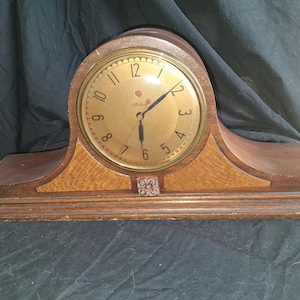 May include: A wooden mantel clock with a curved top and a rectangular base. The clock face is circular, featuring a gold-toned metallic finish with black numerals, thin black hands, and a small decorative floral carving centered below the dial.