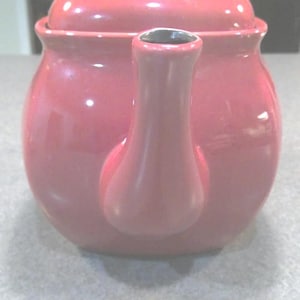 May include: A glossy, red teapot with a matching lid and a black knob. The teapot has a rounded body and a curved spout. The teapot is sitting on a light gray surface.