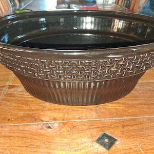 May include: A large, oval-shaped dark brown ceramic slow cooker insert with a glossy finish. The exterior features a textured basket-weave pattern around the upper rim, transitioning into vertical fluted ridges along the lower base section.