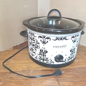 May include: A white Crock-Pot slow cooker with a black damask pattern, a black lid with a handle, and black side handles. The front features the text Crock-Pot, a black control knob with settings for off, low, high, and warm, and a black power cord.